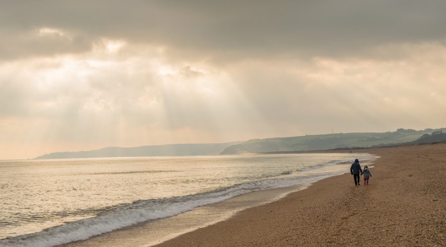 Slapton Sands on a cloudy day with sun beams shinning through the clouds and two people walking away from camera on the beach